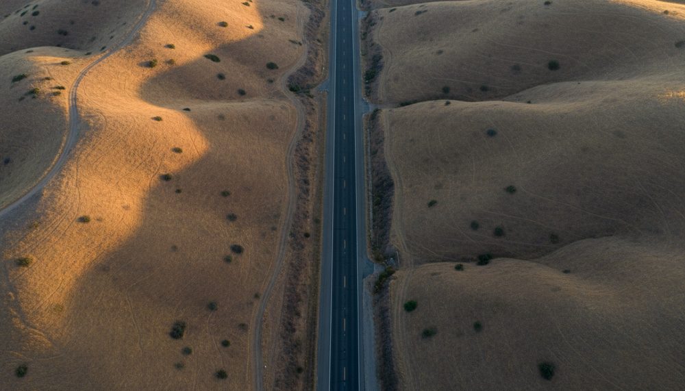 An empty two-lane California highway seen from above, cutting through dry summer hills at dawn.